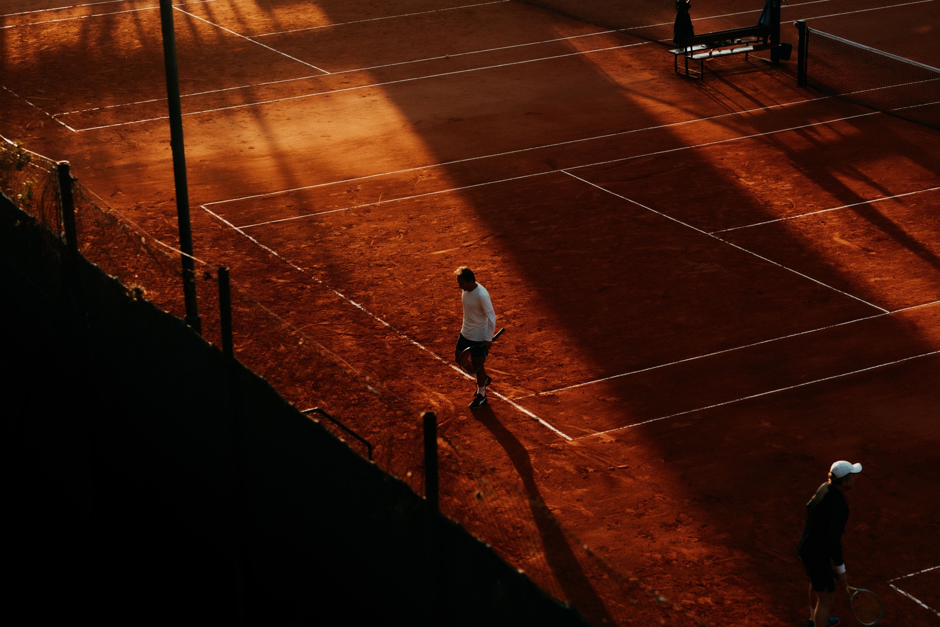 Players on an orange clay tennis court in late afternoon light.